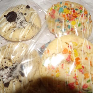 May include: Four cookies in clear packaging. Two cookies have colorful cereal pieces, and white icing drizzle. The other two cookies have chocolate cookie pieces and white icing drizzle. The cookies are round and appear to be soft-baked.