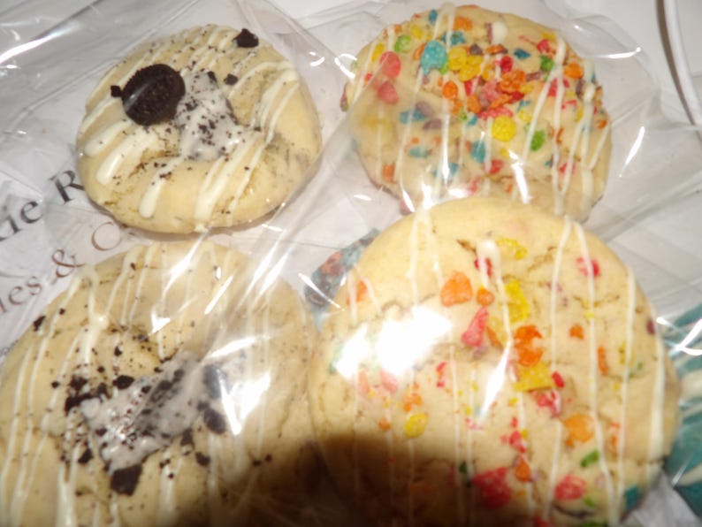 May include: Four cookies in clear packaging. Two cookies have colorful cereal pieces, and white icing drizzle. The other two cookies have chocolate cookie pieces and white icing drizzle. The cookies are round and appear to be soft-baked.