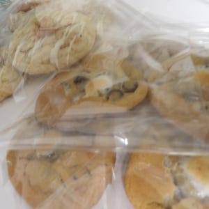 May include: Several baked cookies in clear plastic bags. The cookies have a golden-brown color with various toppings, including what appears to be white icing and dark chocolate chips. The cookies are arranged in a slightly overlapping manner.