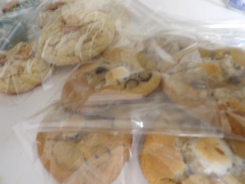 May include: Several baked cookies in clear plastic bags. The cookies have a golden-brown color with various toppings, including what appears to be white icing and dark chocolate chips. The cookies are arranged in a slightly overlapping manner.