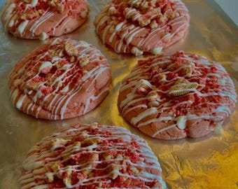 May include: Five pink cookies on a baking sheet. Each cookie is round and topped with white icing drizzled across the top. The cookies are also sprinkled with red and tan cookie pieces.