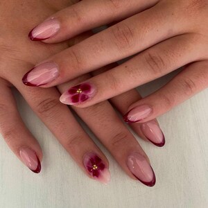Könnte beinhalten: Nahaufnahme von manikürten Nägeln mit French-Tip-Design. Die Nägel haben eine klare Basis mit einer tiefroten Spitze. Zwei Nägel weisen ein Blumendesign mit burgunderfarbenen Blütenblättern und goldenen Akzenten auf. Die Nägel sind oval geformt.