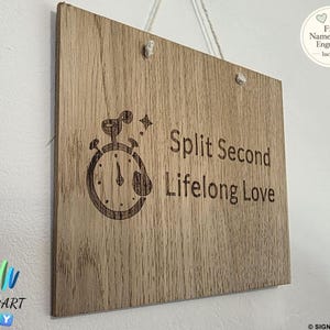 May include: A light brown wooden sign with the words "Split Second Lifelong Love" and a clock illustration. The sign has a wood grain texture and is suspended by a white string. The sign also includes the text "Free Name & Back Engraving Included".