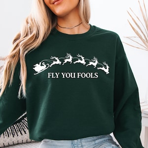 May include: Dark green sweatshirt featuring a white graphic of Santa's sleigh pulled by reindeer. The text "FLY YOU FOOLS" is below the graphic. The sweatshirt is a crew neck style.