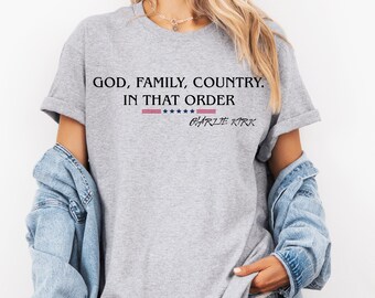 May include: A light gray t-shirt with the text "GOD, FAMILY, COUNTRY. IN THAT ORDER" in black. Below the text is a red and blue stripe design with stars, and the name "CHARLIE KIRK" in black. The shirt is worn with a denim jacket.