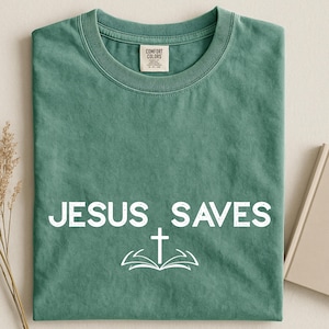 May include: A folded, sage green t-shirt with the words "JESUS SAVES" printed in white, along with a white cross and open book graphic. The shirt has a crew neck and a tag that reads "COMFORT COLORS".