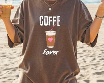 以下が含まれることがあります： 「COFFE lover」の白文字が入った茶色のTシャツ。ピンクのハートが付いたコーヒーカップのグラフィックが「COFFE」の下に配置されています。このシャツは半袖で、ゆったりとしたフィット感です。