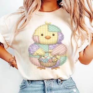 May include: A light beige t-shirt featuring a colorful patchwork chick holding a basket of flowers and Easter eggs. The chick has a yellow gingham head, purple floral wings, and pink striped accents. The basket is woven.