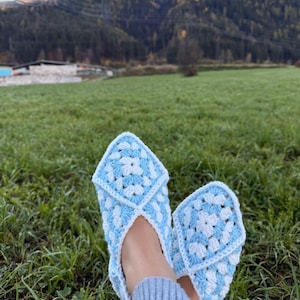 May include: A pair of handmade crochet slippers in a blue and white granny square pattern. The slippers are shown outdoors on a bed of green grass, with a mountain range in the background.