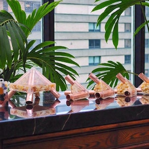 May include: A collection of decorative items, including a pyramid-shaped object and several small, copper-colored structures, are displayed on a dark, reflective surface. The objects are arranged near a window with a view of a city and lush green plants.