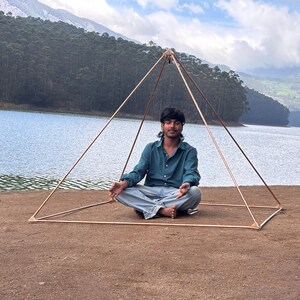 May include: A copper-colored pyramid structure with a person sitting inside in a meditative pose. The pyramid is set up outdoors near a lake and mountains, with a clear sky in the background.