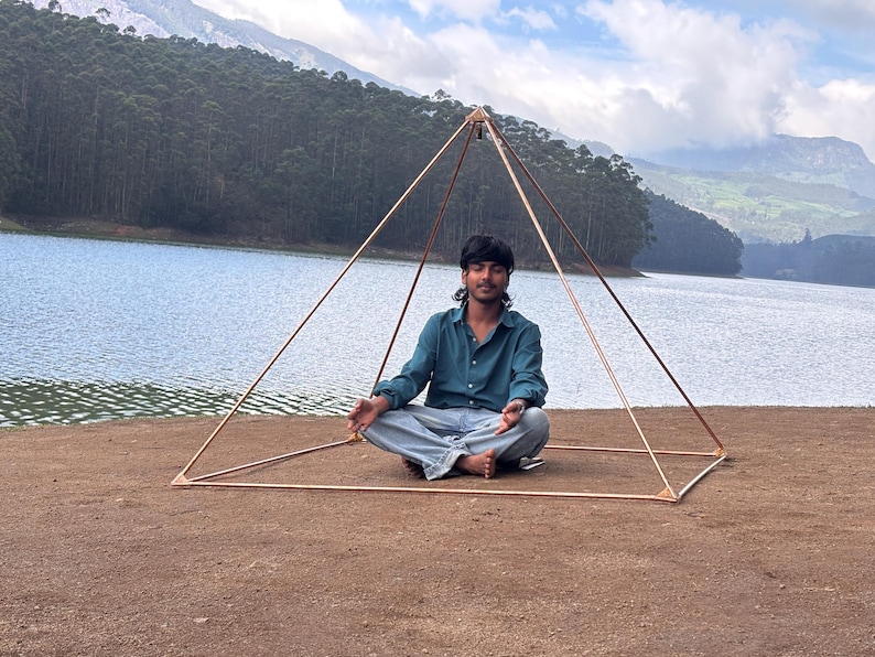 May include: A copper-colored pyramid structure with a person sitting inside in a meditative pose. The pyramid is set up outdoors near a lake and mountains, with a clear sky in the background.