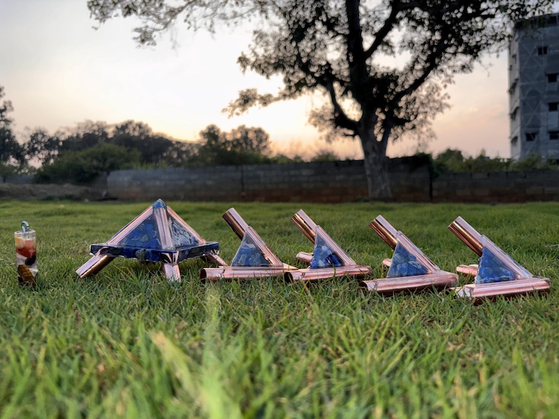 May include: A collection of copper and blue stone pyramids and stands arranged on green grass. The pyramid has a square base and is supported by copper legs. The stands have a copper frame with a blue stone insert.