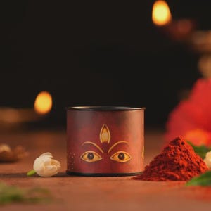 May include: A cylindrical container with a dark red finish featuring a graphic illustration of two eyes and a third eye symbol on the front. The container is positioned next to a small white flower and a pile of vibrant red powder.