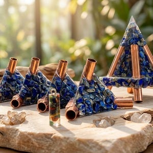 May include: A collection of handcrafted objects featuring blue lapis lazuli stones and copper accents. The items include pyramid-shaped pieces, a small pendant, and other geometric forms, all displayed on a natural stone surface.