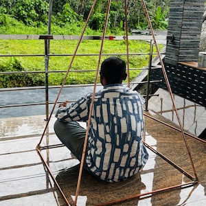 May include: A copper-colored pyramid-shaped structure with a person sitting inside. The pyramid is constructed of thin metal rods, forming a geometric shape. The person is wearing a blue and white patterned shirt, seated on a reflective, brown-toned surface.