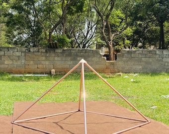 Puede incluir: Una estructura piramidal de metal color cobre, fabricada con tubos huecos y delgados. El objeto presenta una base cuadrada con cuatro lados que se elevan hacia un vértice único en la parte superior, formando una pirámide geométrica.