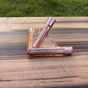 May include: A copper-colored metal object, possibly a decorative piece or a component of a larger structure, is positioned on a wooden surface. The object has a geometric shape, with three connected tubes. The background is a wooden table and green grass.