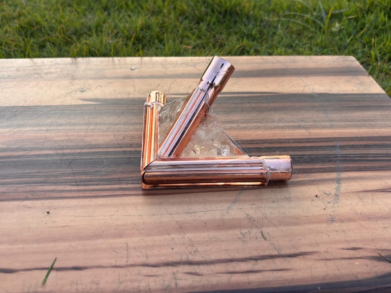 May include: A copper-colored metal object, possibly a decorative piece or a component of a larger structure, is positioned on a wooden surface. The object has a geometric shape, with three connected tubes. The background is a wooden table and green grass.