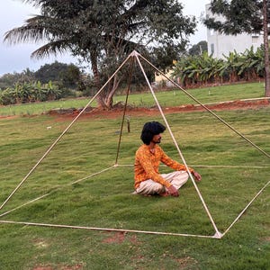 May include: A large, open-frame pyramid structure made of metal tubing, set on a grassy lawn. A person sits inside the pyramid, cross-legged, wearing an orange patterned shirt and light pants. The pyramid is outdoors, with trees and a building in the background.