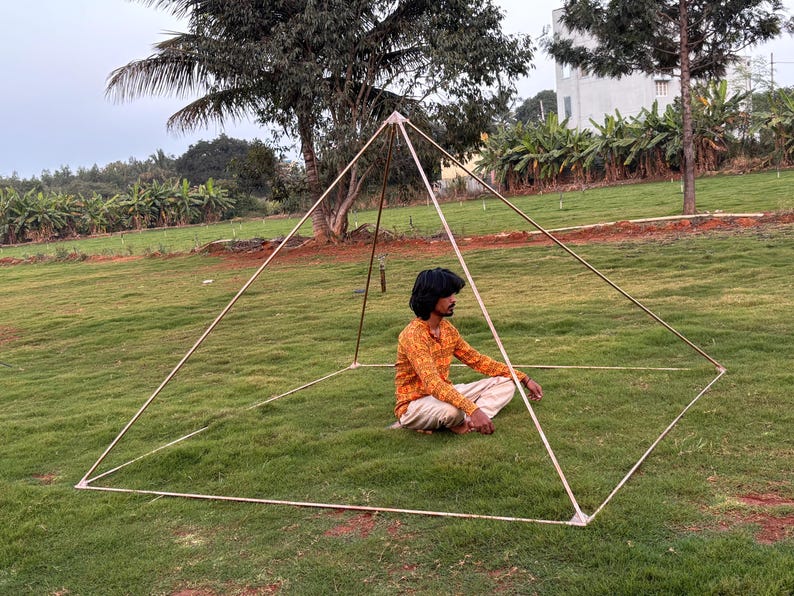 May include: A large, open-frame pyramid structure made of metal tubing, set on a grassy lawn. A person sits inside the pyramid, cross-legged, wearing an orange patterned shirt and light pants. The pyramid is outdoors, with trees and a building in the background.
