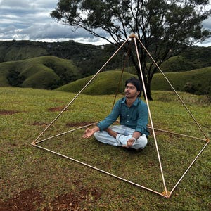May include: A copper-colored pyramid structure with a person meditating inside, set on a grassy hill with a tree and rolling green hills in the background. The pyramid is made of thin rods joined at the corners.
