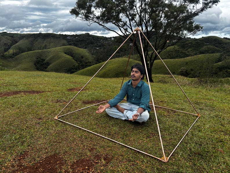 May include: A copper-colored pyramid structure with a person meditating inside, set on a grassy hill with a tree and rolling green hills in the background. The pyramid is made of thin rods joined at the corners.