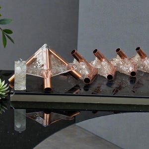 May include: A collection of copper and crystal decorative objects. The centerpiece is a pyramid shape with copper tubing and clear crystals. Adjacent are four copper tubes with crystal accents. A clear, cylindrical object is also present.