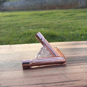 May include: A copper-colored, right-angled pipe fitting, possibly for plumbing or decorative use. The fitting is made of shiny metal and is positioned on a wooden surface. The background features green grass and a blurred view of a fence.