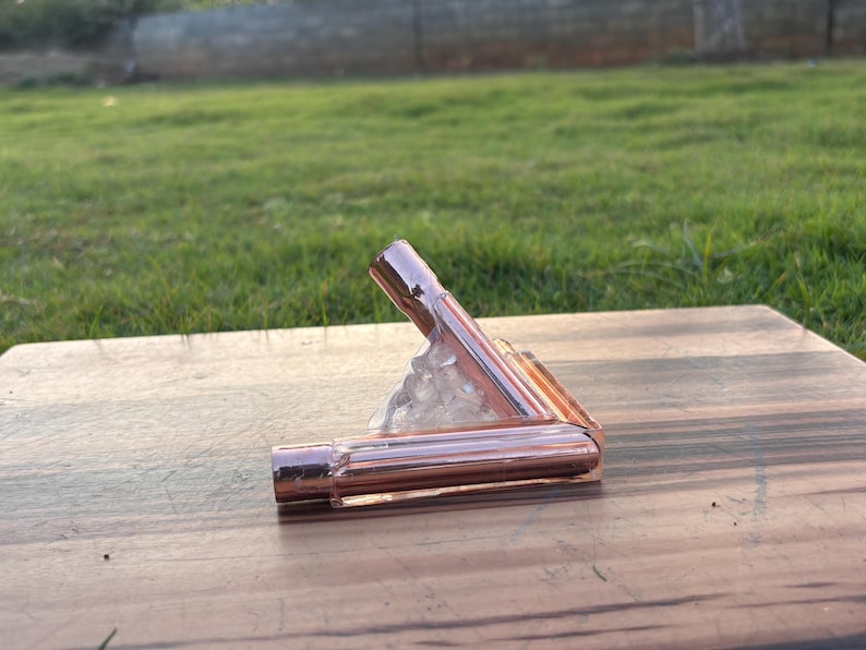 May include: A copper-colored, right-angled pipe fitting, possibly for plumbing or decorative use. The fitting is made of shiny metal and is positioned on a wooden surface. The background features green grass and a blurred view of a fence.