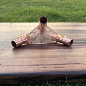 May include: A copper pipe fitting with three openings, set in a clear, hardened resin. The fitting is positioned on a wooden surface with a natural wood grain pattern. The background features green grass.