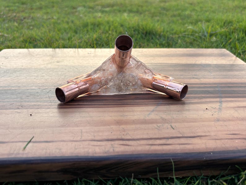 May include: A copper pipe fitting with three openings, set in a clear, hardened resin. The fitting is positioned on a wooden surface with a natural wood grain pattern. The background features green grass.