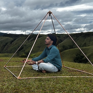 May include: A copper-colored pyramid structure with a person meditating inside. The pyramid is set on a grassy hill with a backdrop of rolling green hills and a cloudy sky. The person is wearing a blue shirt and white pants.