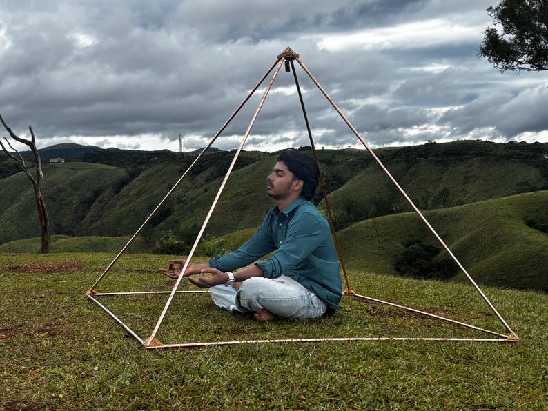 May include: A copper-colored pyramid structure with a person meditating inside. The pyramid is set on a grassy hill with a backdrop of rolling green hills and a cloudy sky. The person is wearing a blue shirt and white pants.