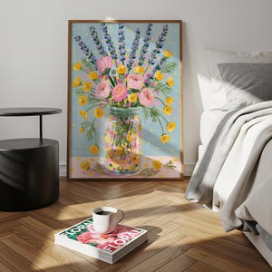 May include: A framed floral art print featuring a bouquet of pink and yellow flowers and lavender sprigs in a mason jar. The artwork is set against a light blue background with a pink border. A coffee cup sits on a magazine.