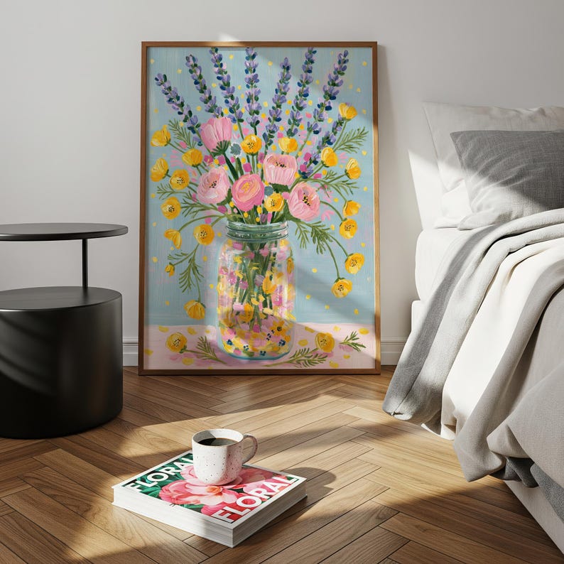 May include: A framed floral art print featuring a bouquet of pink and yellow flowers and lavender sprigs in a mason jar. The artwork is set against a light blue background with a pink border. A coffee cup sits on a magazine.