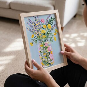 May include: A framed floral print featuring a mason jar filled with colorful flowers. The artwork showcases yellow, pink, and purple blooms against a light blue background. The frame is made of light-colored wood.