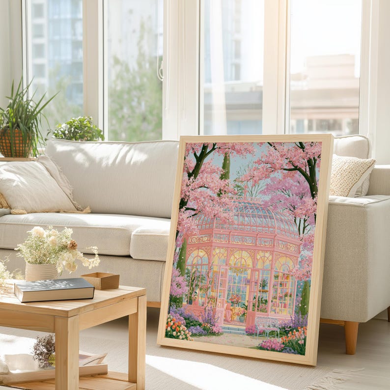 May include: A framed art print featuring a pink greenhouse scene with cherry blossom trees. The artwork has a light wooden frame and is leaning against a white sofa in a well-lit living room. The print showcases a vibrant garden.