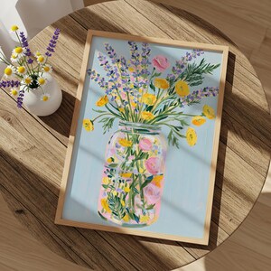 May include: A framed floral art print featuring a vibrant bouquet of lavender, yellow, and pink flowers in a mason jar against a light blue background. The artwork is displayed on a round wooden table, with a small vase of flowers nearby.