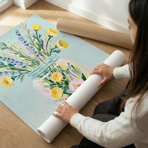May include: A floral art print featuring a jar filled with yellow and pink roses, lavender, and other flowers. The artwork is on a light blue background. A person is holding a rolled-up white print.