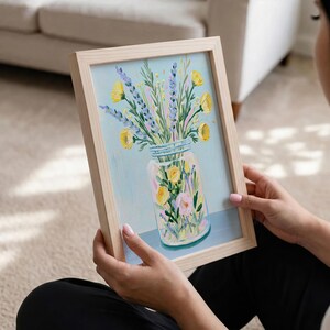 May include: A framed floral painting featuring a mason jar filled with yellow and pink flowers, and purple lavender. The artwork has a light blue background and is held by a person.