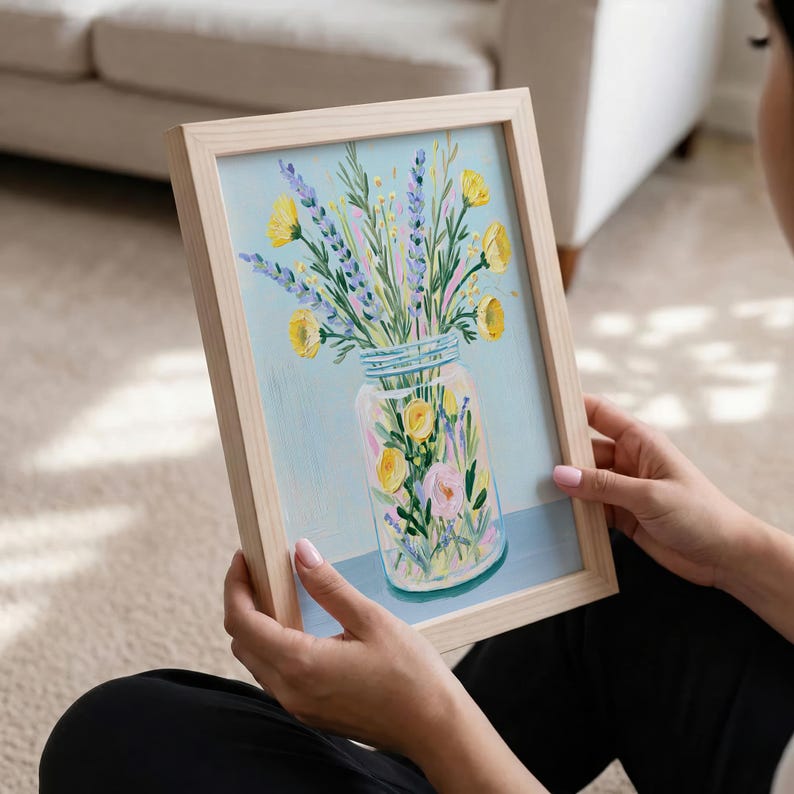 May include: A framed floral painting featuring a mason jar filled with yellow and pink flowers, and purple lavender. The artwork has a light blue background and is held by a person.