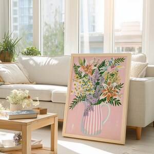 Pu&ograve; includere: Stampa floreale incorniciata con un bouquet di gigli, margherite e lavanda in una brocca a righe rosa e bianche. L'opera d'arte ha una cornice in legno chiaro ed &egrave; ambientata su uno sfondo rosa. La stampa &egrave; esposta in un soggiorno.