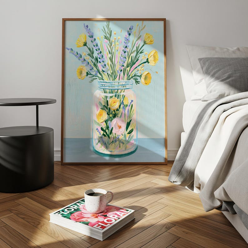 May include: A framed floral art print featuring a glass jar filled with yellow, pink, and purple flowers. The artwork has a light blue background and a wooden frame. A coffee cup sits on a magazine with the word "FLORAL".