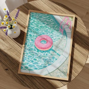 Puede incluir: Una lámina artística rectangular enmarcada que muestra una piscina de agua turquesa, un flotador rosa con forma de dónut y una escalera de metal rosa. La obra está expuesta sobre una mesa redonda de madera junto a un jarrón blanco con flores.