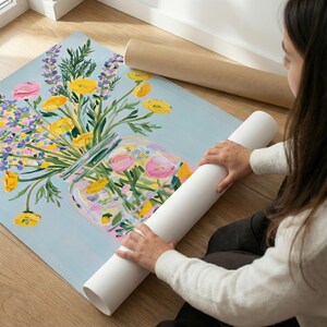 May include: A floral painting featuring a bouquet of yellow, pink, and purple flowers in a glass jar against a light blue background. Two rolled-up paper tubes are visible. The artwork is on a wooden floor.