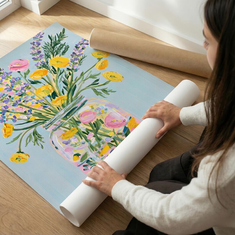 May include: A floral painting featuring a bouquet of yellow, pink, and purple flowers in a glass jar against a light blue background. Two rolled-up paper tubes are visible. The artwork is on a wooden floor.