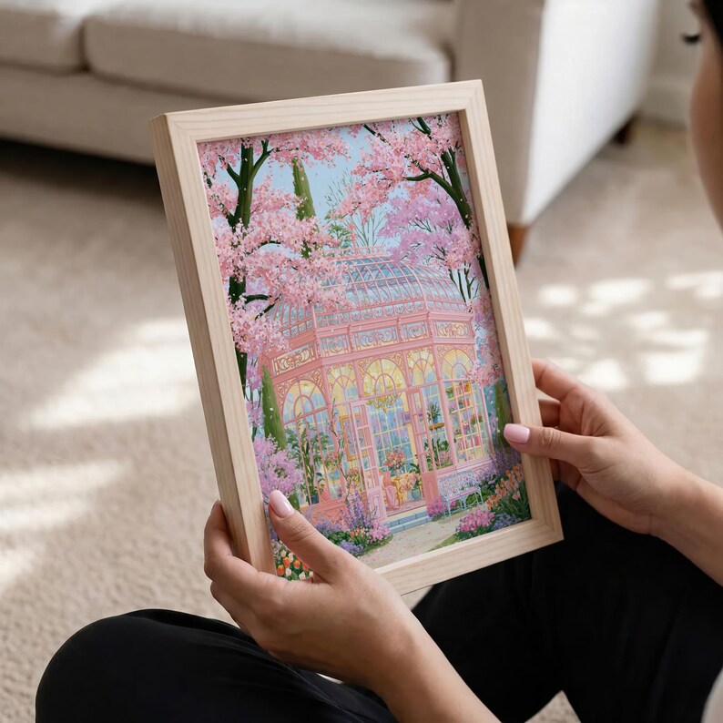 May include: A framed print featuring a pink greenhouse surrounded by cherry blossom trees. The artwork has a light wood frame and is held by a person. The greenhouse has arched windows and is filled with plants.