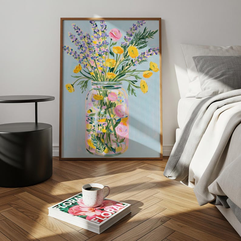May include: A framed floral art print featuring a jar filled with colorful flowers. The artwork showcases yellow, pink, and purple blooms against a light blue background. The print is displayed on a wooden floor, with a coffee table and a magazine nearby.