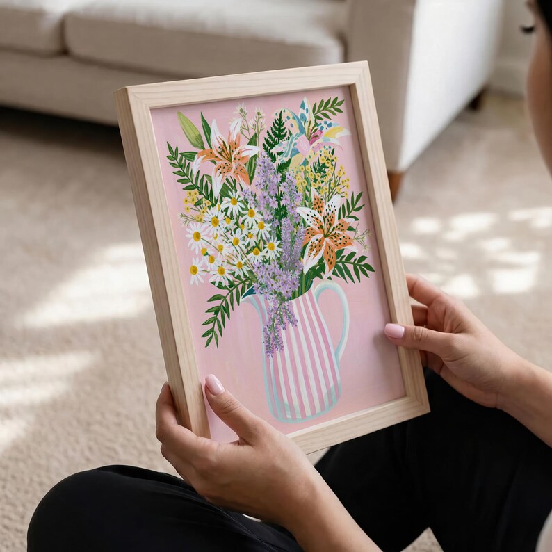 Pu&ograve; includere: Una stampa floreale incorniciata con un bouquet di gigli, margherite e lavanda in una brocca a righe. L'opera d'arte ha uno sfondo rosa e una cornice in legno chiaro. L'immagine &egrave; tenuta da una persona.